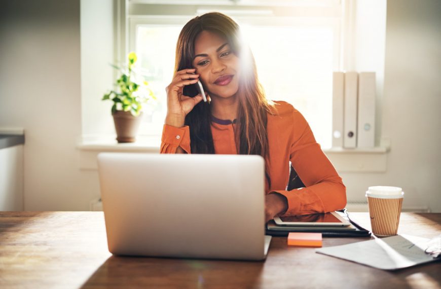 Smiling young African female entrepreneur talking on a cellphone and working on a laptop while sitting at a table in her home office